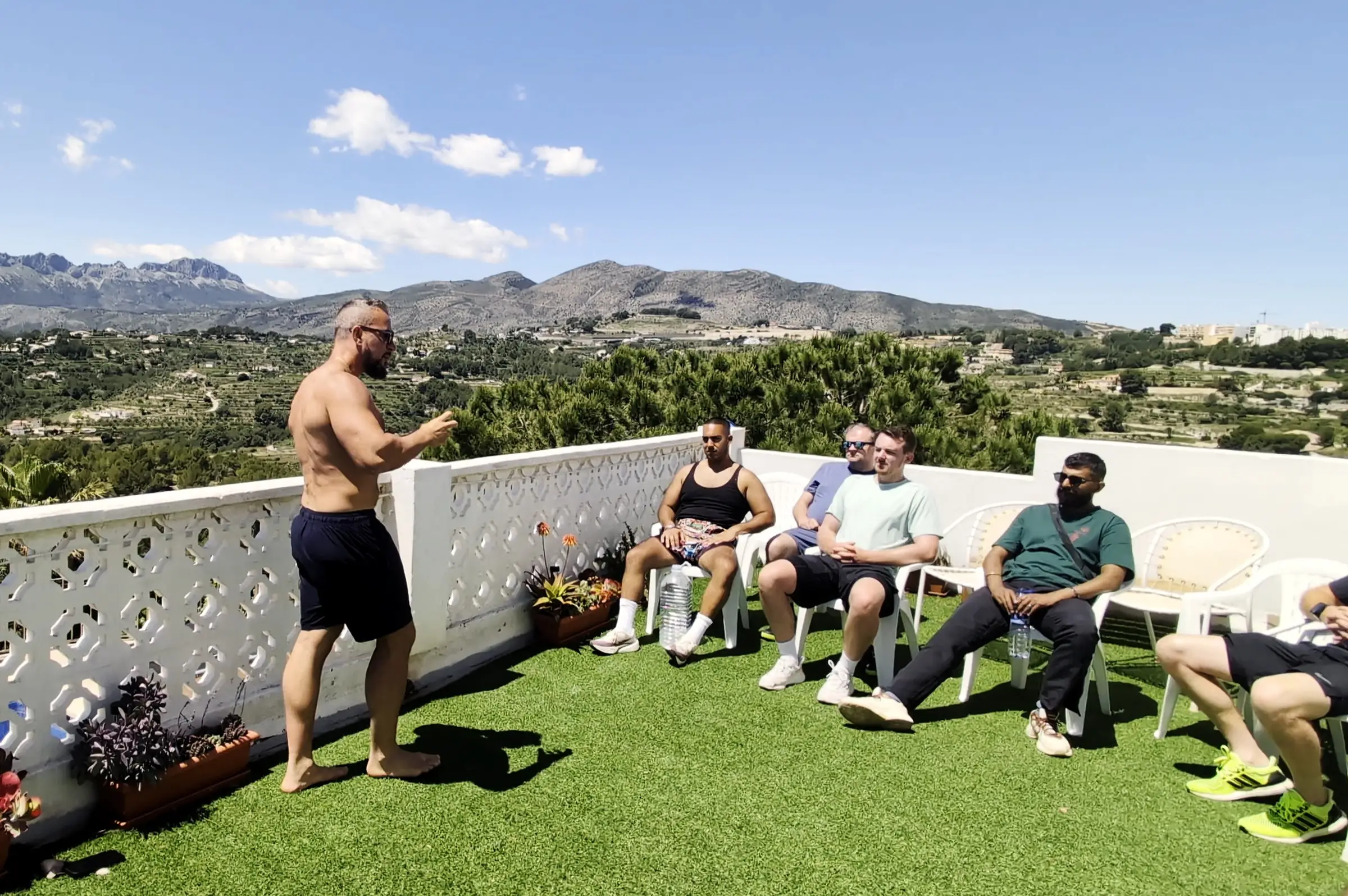 Steve Walker on the villa roof terrace, talking to the seated group, mountains behind