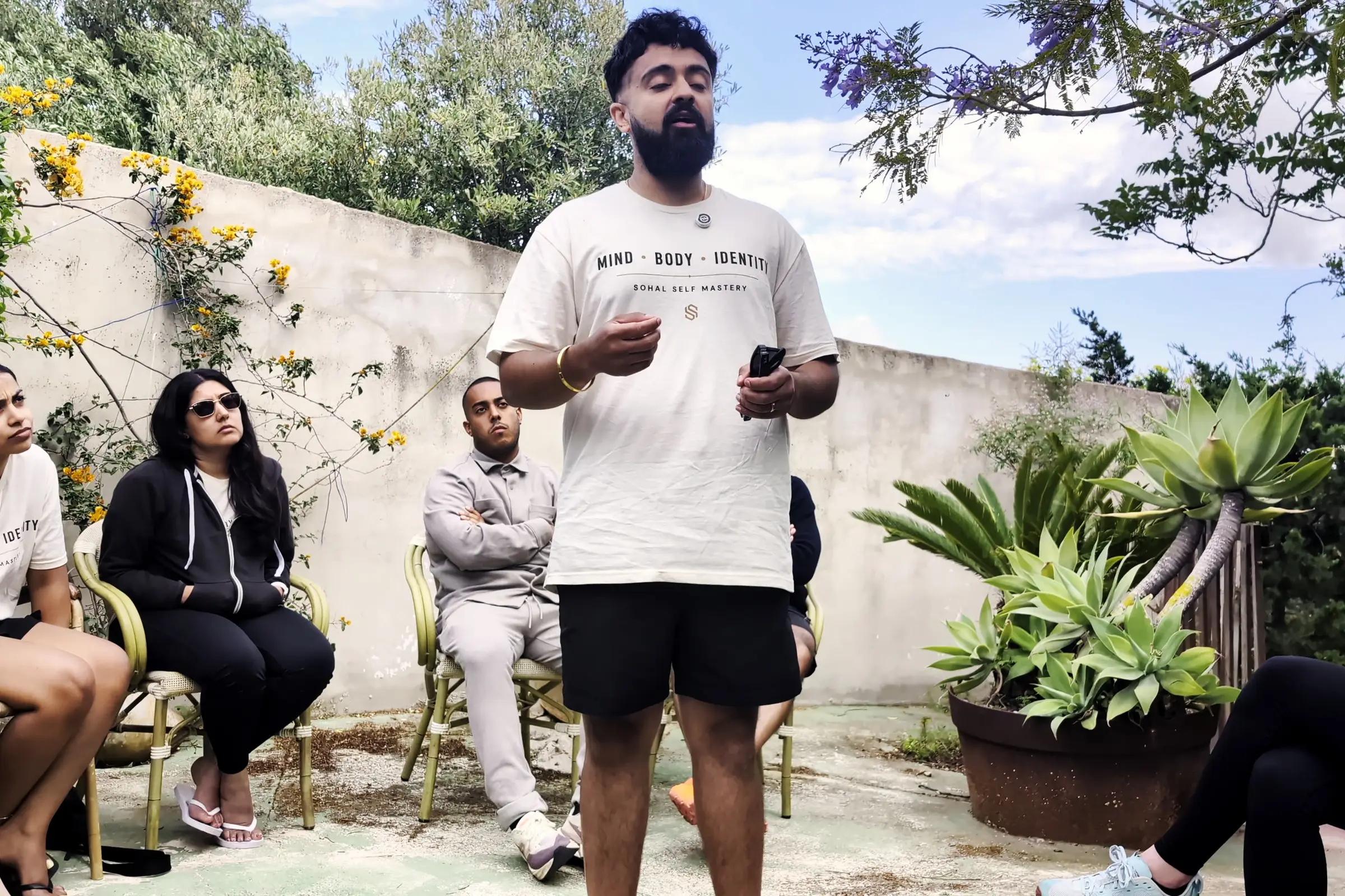 Sukh Sohal standing in a villa garden in the team T-shirt, talking to the group seated round him